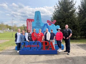 A group of people stands outdoors in front of a large colorful "America 250 PA" display featuring Pennsylvania-themed illustrations, with trees and a grassy field in the background. A police officer stands at the far right.