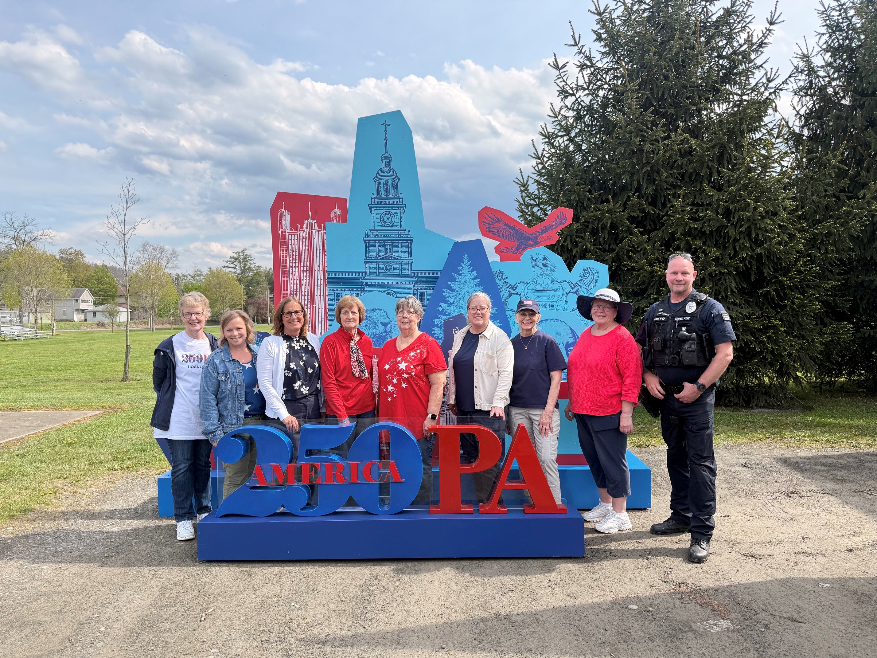 A group of people stands outdoors in front of a large colorful "America 250 PA" display featuring Pennsylvania-themed illustrations, with trees and a grassy field in the background. A police officer stands at the far right.
