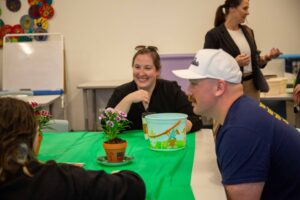 Several people sit around a table covered with a green tablecloth, working with small potted flowers and a decorative bucket in a classroom setting. Shelves, colorful art pieces, and tables are visible in the background.