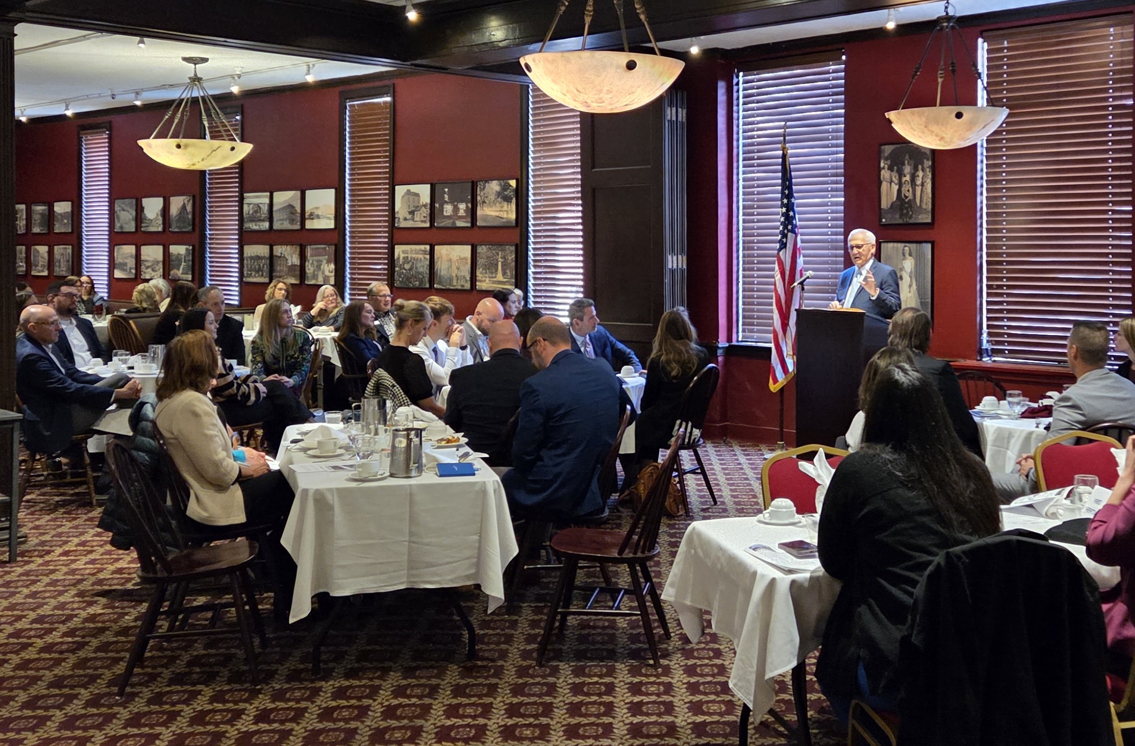 Sen. Yaw addressing a seated audience in a formal dining room with framed historical photos on the walls and an American flag positioned beside the podium.