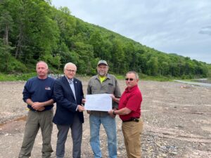 Sen. Gene Yaw and three people stand together outdoors on a gravel-covered site surrounded by dense green forested hills. They are holding a large set of printed plans or drawings.