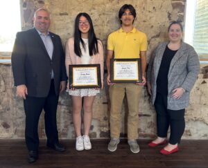 Two students stand in the center holding framed certificates, accompanied by two adults on each side, posing indoors against a rustic stone wall.