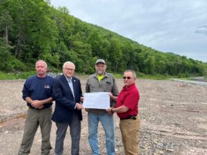 Sen. Gene Yaw stands with Lewis Township officials and emergency management representatives on a large gravel lot surrounded by wooded mountainsides in Trout Run. The group holds a large rendering of the planned municipal building site.