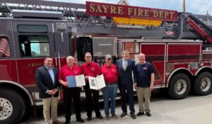 Six people stand in front of a red fire truck labeled “Sayre Fire Dept.” Two of them hold framed proclamations or certificates.