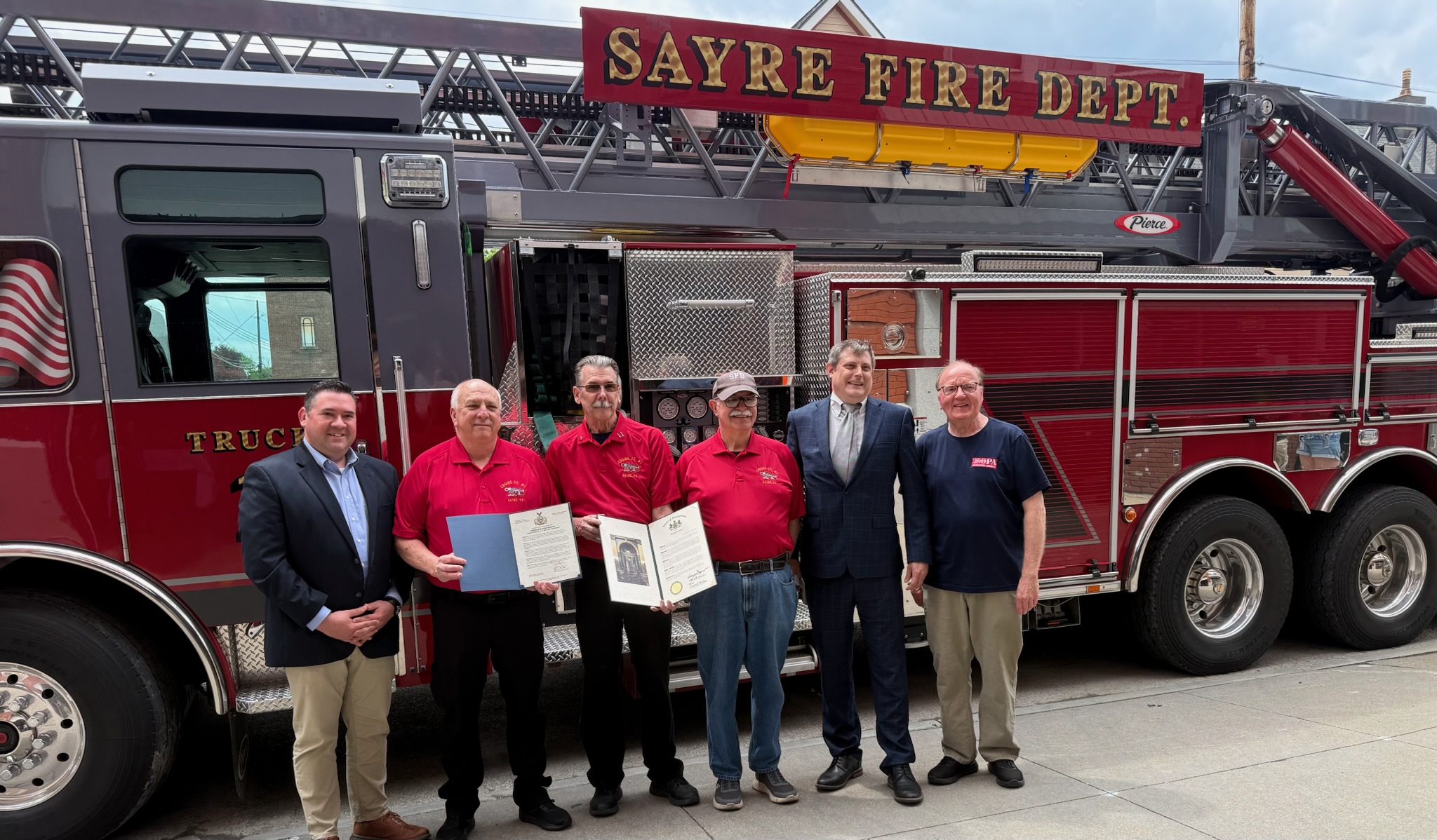 Six people stand in front of a red fire truck labeled “Sayre Fire Dept.” Two of them hold framed proclamations or certificates.
