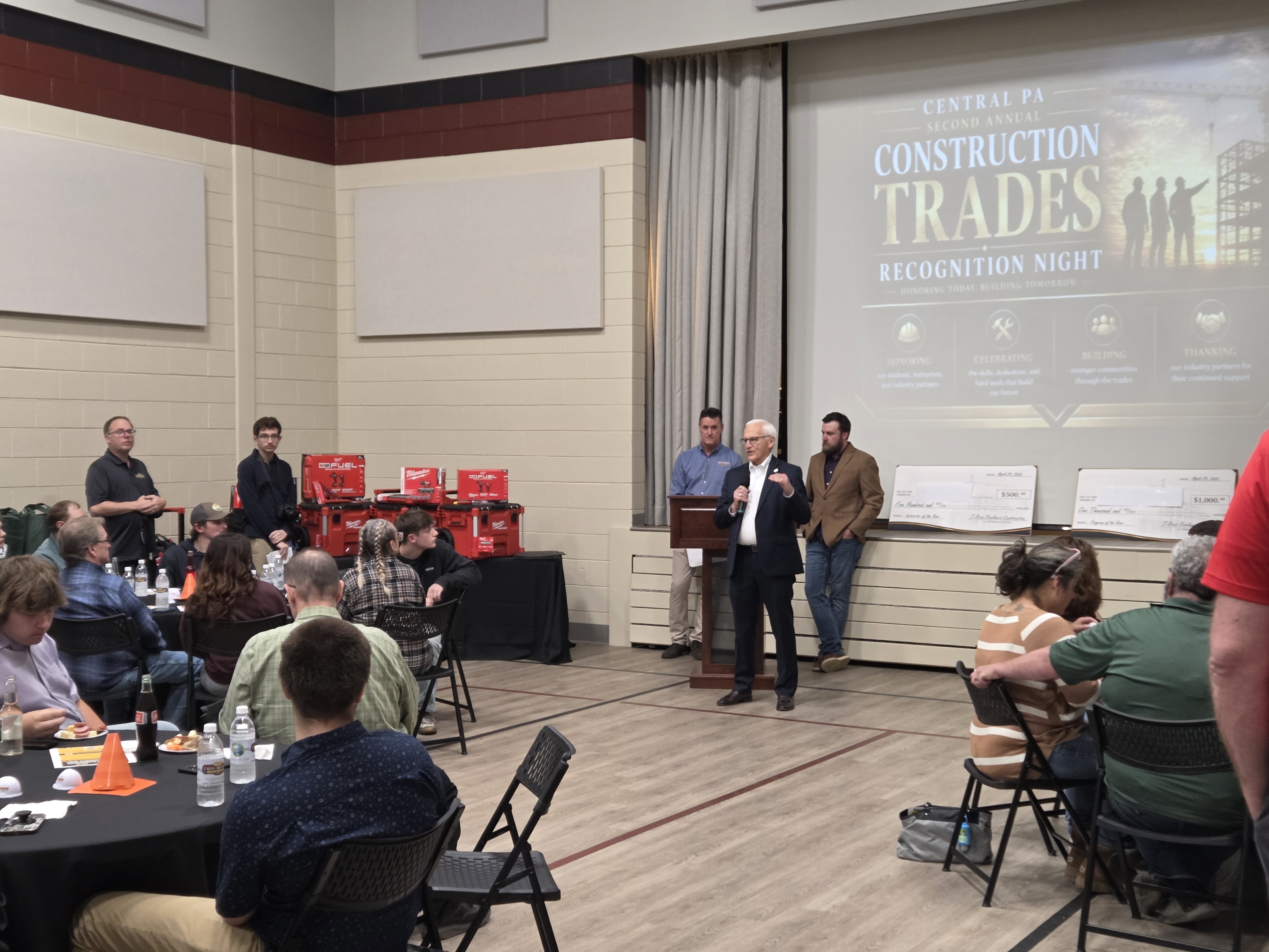 An indoor event with attendees seated at tables while Sen. Yaw addresses the audience from a podium in front of a large screen displaying “Construction Trades Recognition Night.”