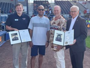 Sen. Yaw and three people standing on a baseball field, with two individuals holding framed certificates. Stadium seating and spectators are visible in the background.