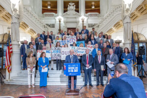 A large group of people stands on the steps inside a grand government building holding signs supporting natural gas and energy jobs, while Yaw stands at a podium with a “Welcome to Natural Gas & Oil Day” sign.
