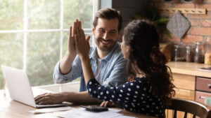 Happy young family couple giving high five to each other, finishing managing household budget, celebrating successful investment, making all payments online in e-banking computer application.