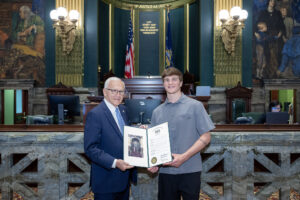Sen. Yaw and Gage Wentzel standing inside a legislative chamber, holding a framed certificate together. The room features marble details, murals, flags, and formal seating.