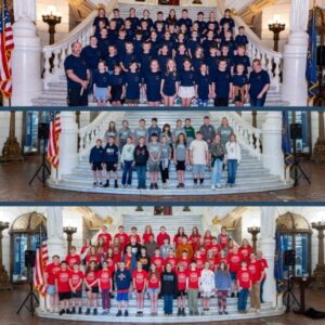 A large group of children and adults standing on the grand marble staircase inside an ornate government building, with statues, flags, and tall lamps in the background.