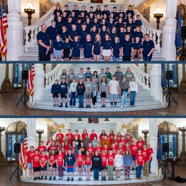 A large group of children and adults standing on the grand marble staircase inside an ornate government building, with statues, flags, and tall lamps in the background.