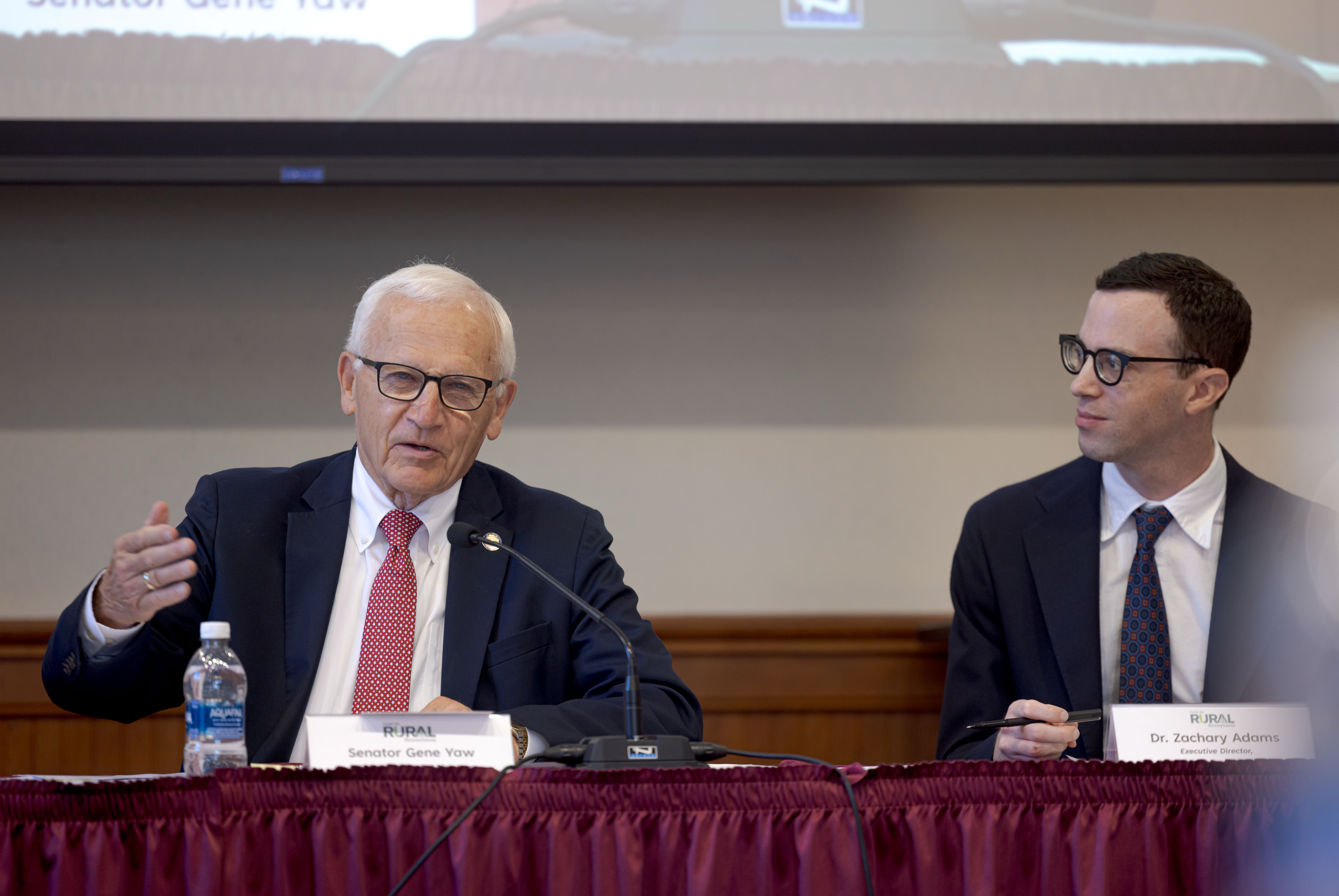 A panel discussion with Sen. Yaw and another person seated at a table. Sen. Yaw is speaking into the microphone while gesturing with one hand. The person on the right looks toward the speaker. Both are dressed in business attire.