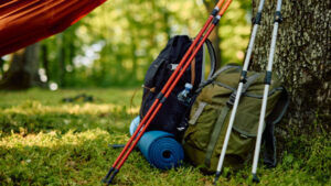 Hiking backpacks, trekking poles, a rolled-up sleeping mat, and a water bottle lie on the grass under a hammock, ready for a hike in nature