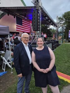 Sen. Yaw and a woman stand on a grassy area in front of a concert stage decorated with an American flag and lighting equipment.