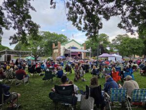 A large crowd sits on lawn chairs facing an outdoor performance stage surrounded by trees in a park setting.