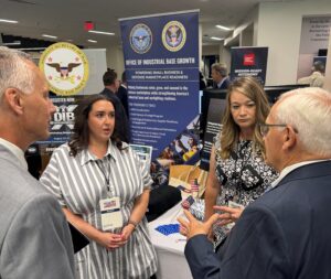 Sen. Gene Yaw and three people stand in discussion near informational display banners related to industrial base growth and defense programs at an indoor event.