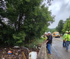 Sen. Yaw and Several workers stand along a roadside bridge, inspecting storm debris piled against the edge while a utility truck is parked nearby.