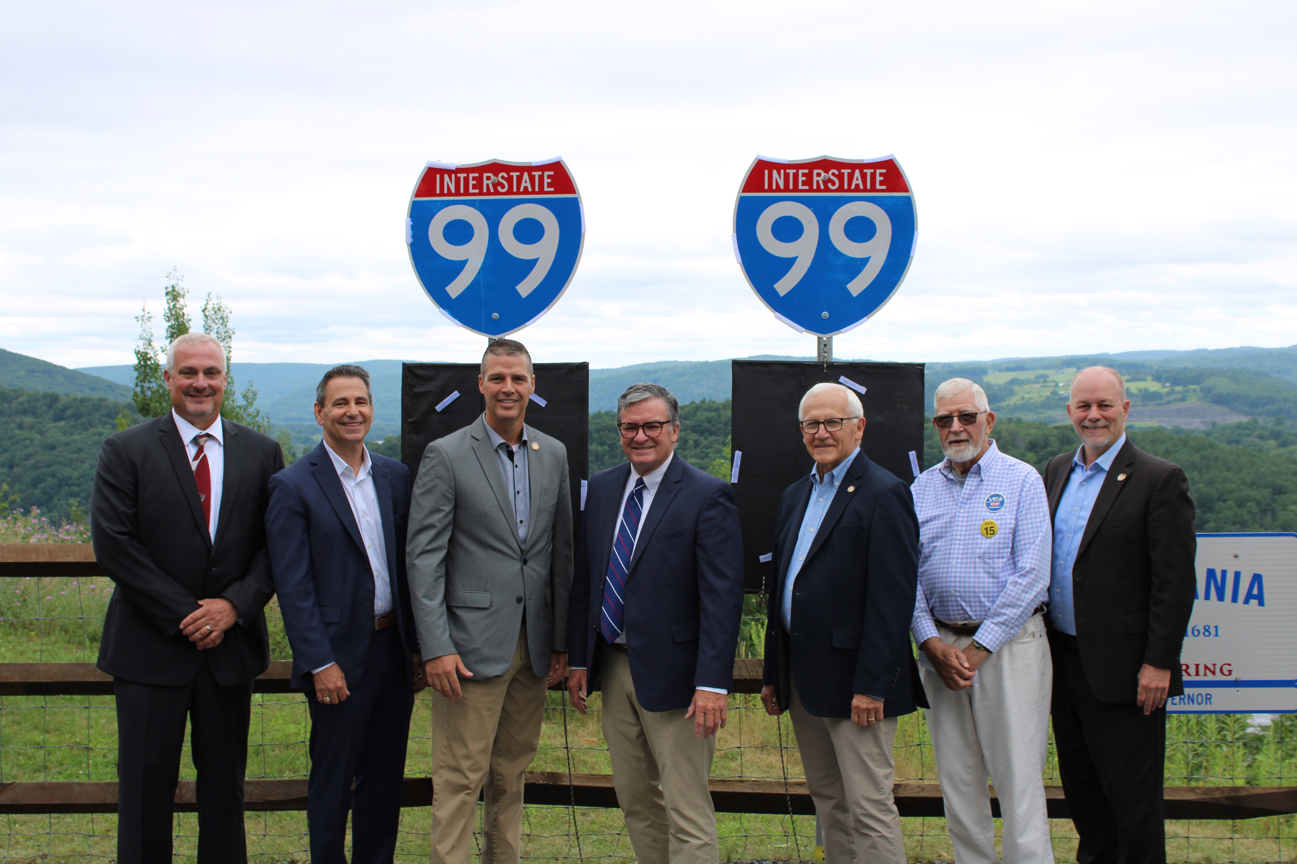 Sen. Yaw and six people standing side by side outdoors in front of two Interstate 99 highway signs. Rolling green hills and a cloudy sky appear in the background.