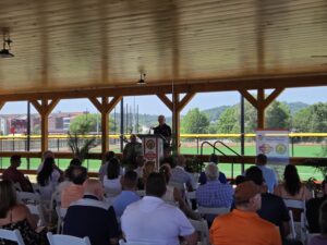 An audience sits under a wooden pavilion while Sen. Yaw addresses them from a podium, with a baseball field visible behind the structure.