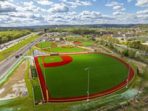 Aerial view of multiple bright green and red baseball fields near a highway, with surrounding buildings and rolling hills in the background.