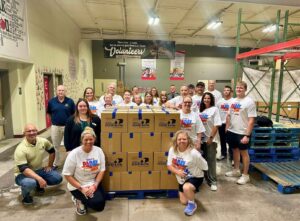 A large volunteer group poses inside a food bank warehouse behind a pallet stacked with boxes labeled “Central PA Food Bank.”