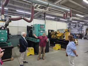 Sen. Gene Yaw and Sen. Lynda Schlegel Culver are joined by a person in a workshop explaining machinery operations while standing between large industrial generators and ventilation tubing overhead.