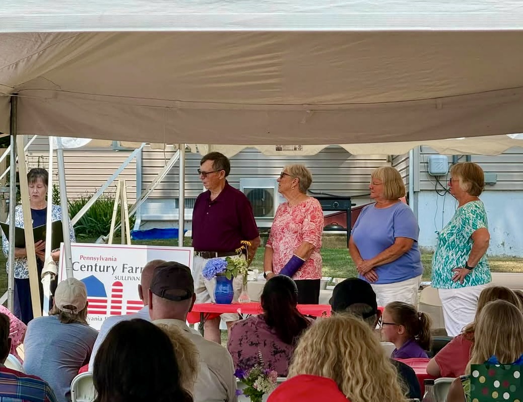 Several people standing under a large event tent while an audience sits watching. A sign reading “Pennsylvania Century Farm” is displayed near the front of the tent.