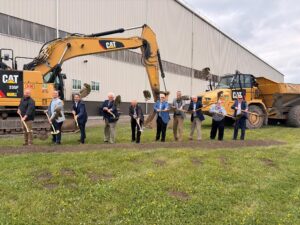 Sen. Yaw and A group of people standing in a row outdoors, each holding a shovel and lifting dirt during a ceremonial groundbreaking. Large construction equipment is behind them.
