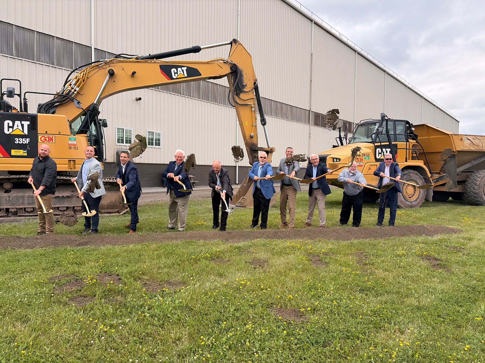 Sen. Yaw and A group of people standing in a row outdoors, each holding a shovel and lifting dirt during a ceremonial groundbreaking. Large construction equipment is behind them.