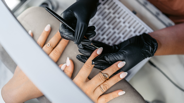Close up of pair of hands with white nails receiving a manicure from second pair of hands in black gloves.]