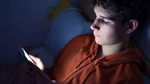 Young male in orange hooded sweatshirt on bed in the dark staring at cellphone.