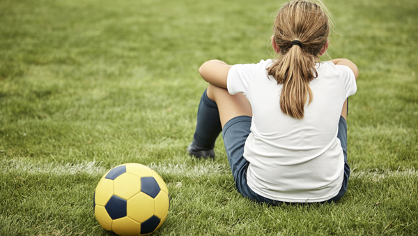 Rear view of girl by soccer ball in field. Female player sitting in training ground. She is in sportswear.