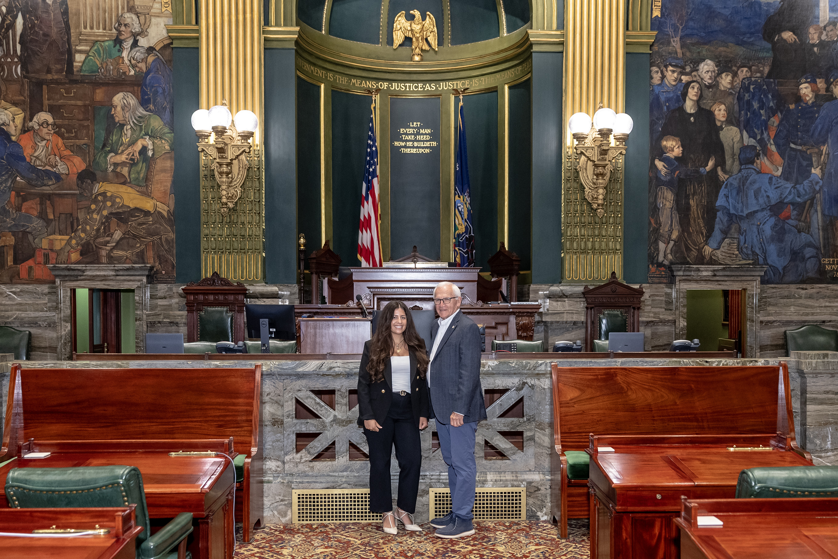 Yaw and a woman stand inside a legislative chamber, posing for a photo in front of a judge’s bench and ornate murals.