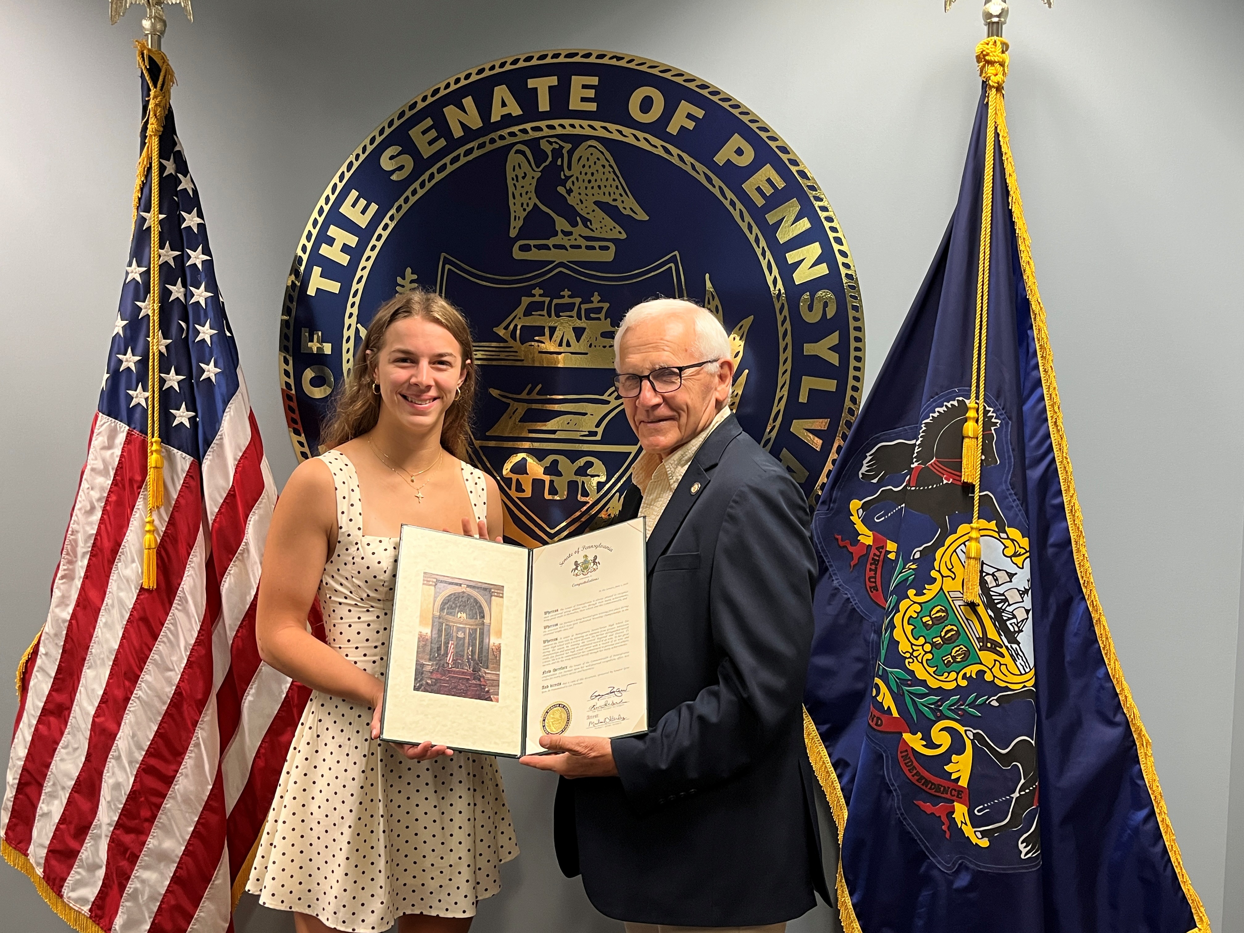 Yaw and a woman stand indoors in front of a large state government seal and two flags, holding an official certificate together.