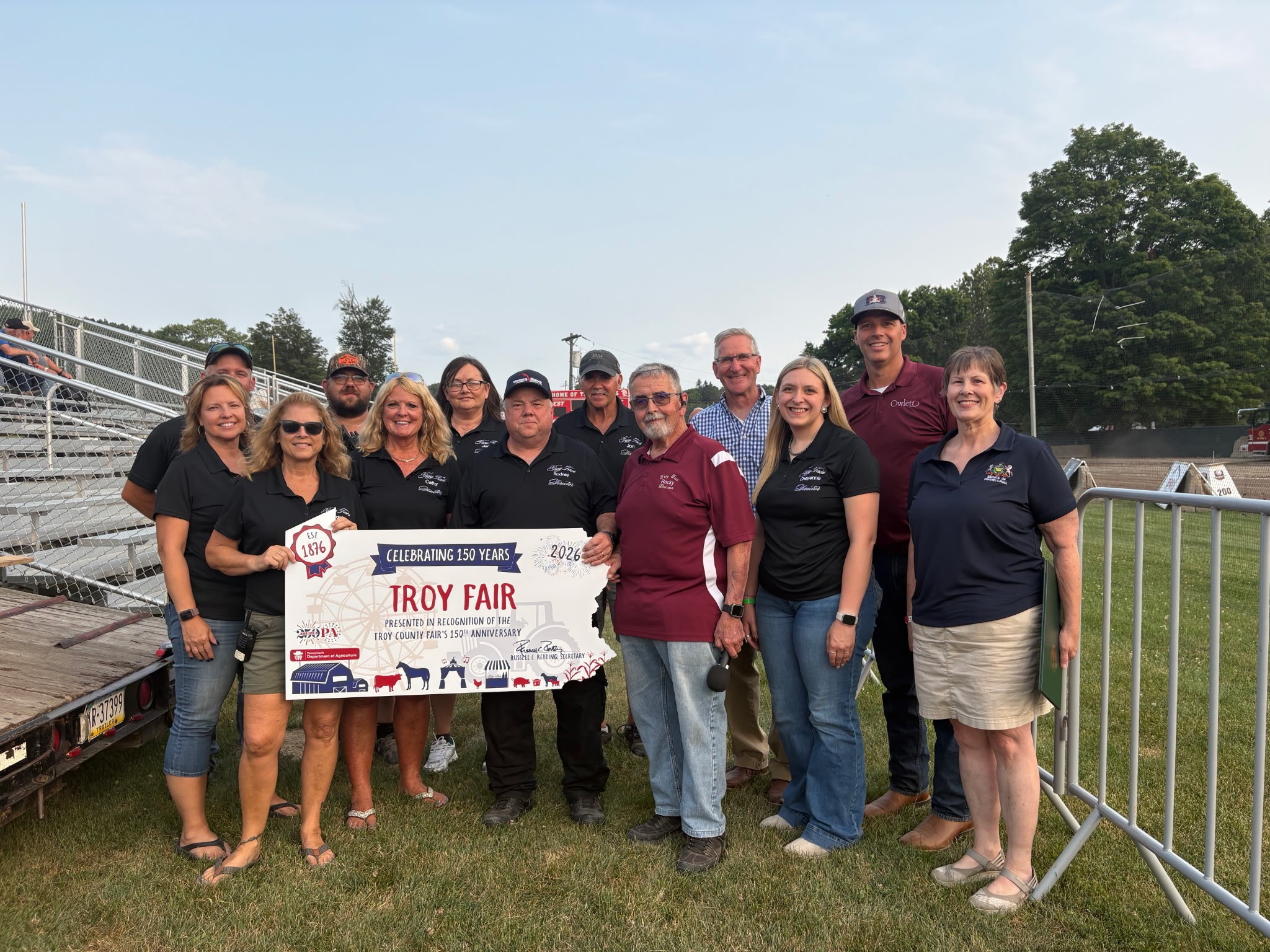 A group of people standing outdoors on grass near bleachers, holding a large sign that celebrates the 150th anniversary of the Troy Fair. The group is smiling and posing for the photo.