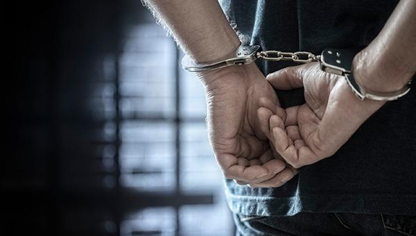 Closeup of standing man’s hands handcuffed behind his back with shadows in background.