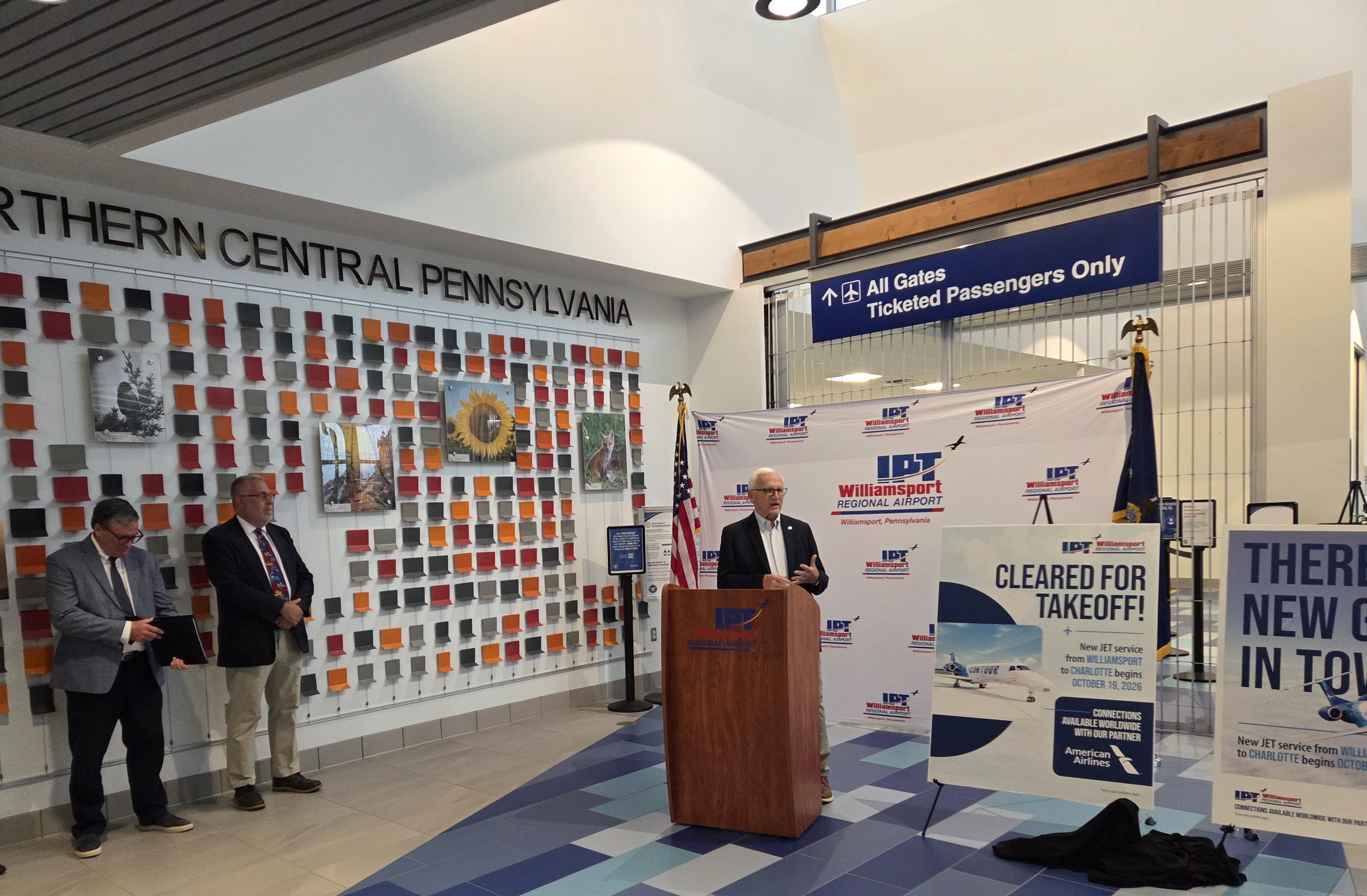 A group of adults standing inside an airport terminal near a podium with signage for a regional airport. Yaw is at the podium, while others stand to the side. Behind them are displays with regional photography and flight announcement posters.