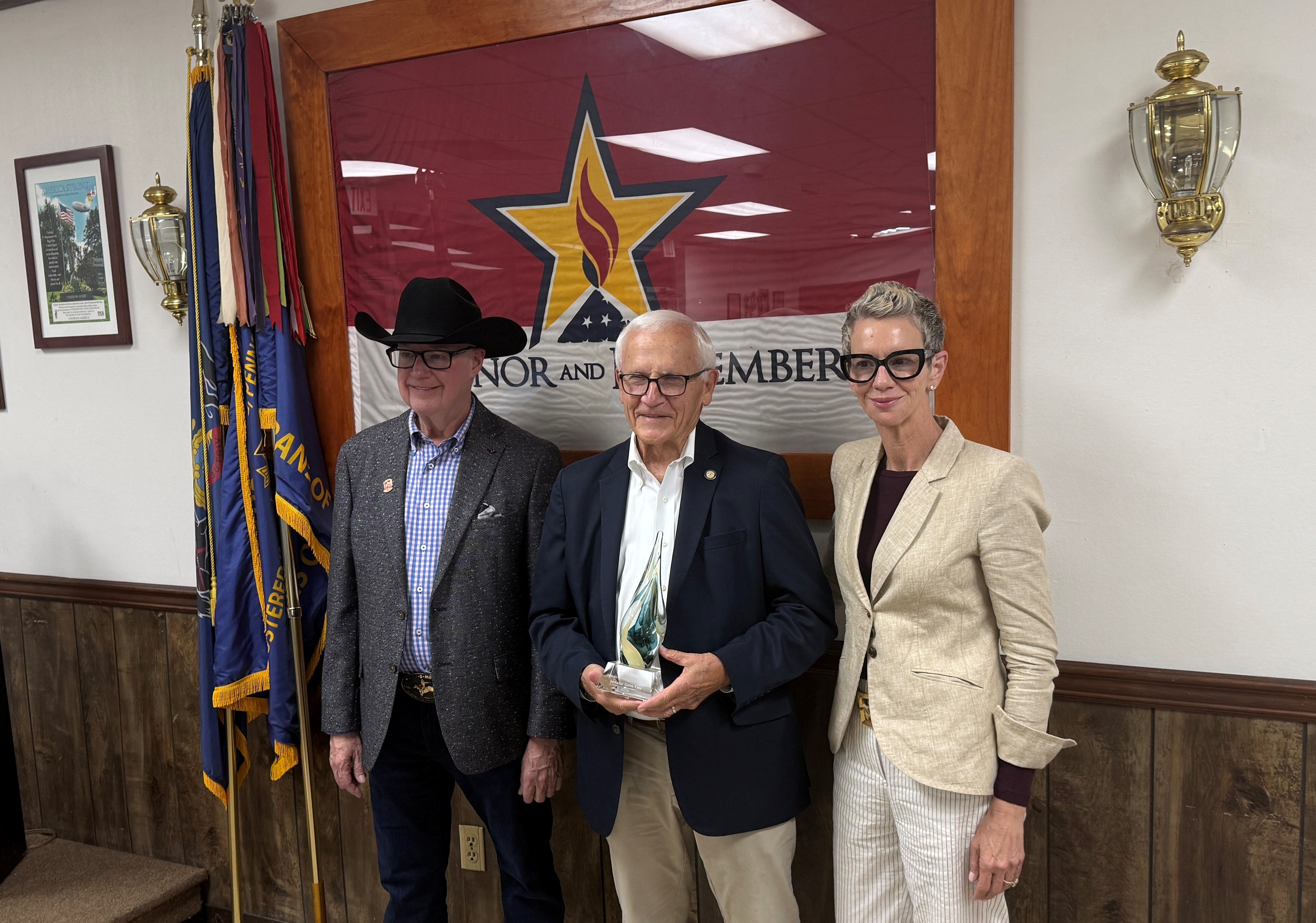 Sen. Yaw and two adults stand together posing for a photo in front of a large banner with a star emblem. Yaw holds a glass award, and flags are displayed beside them.