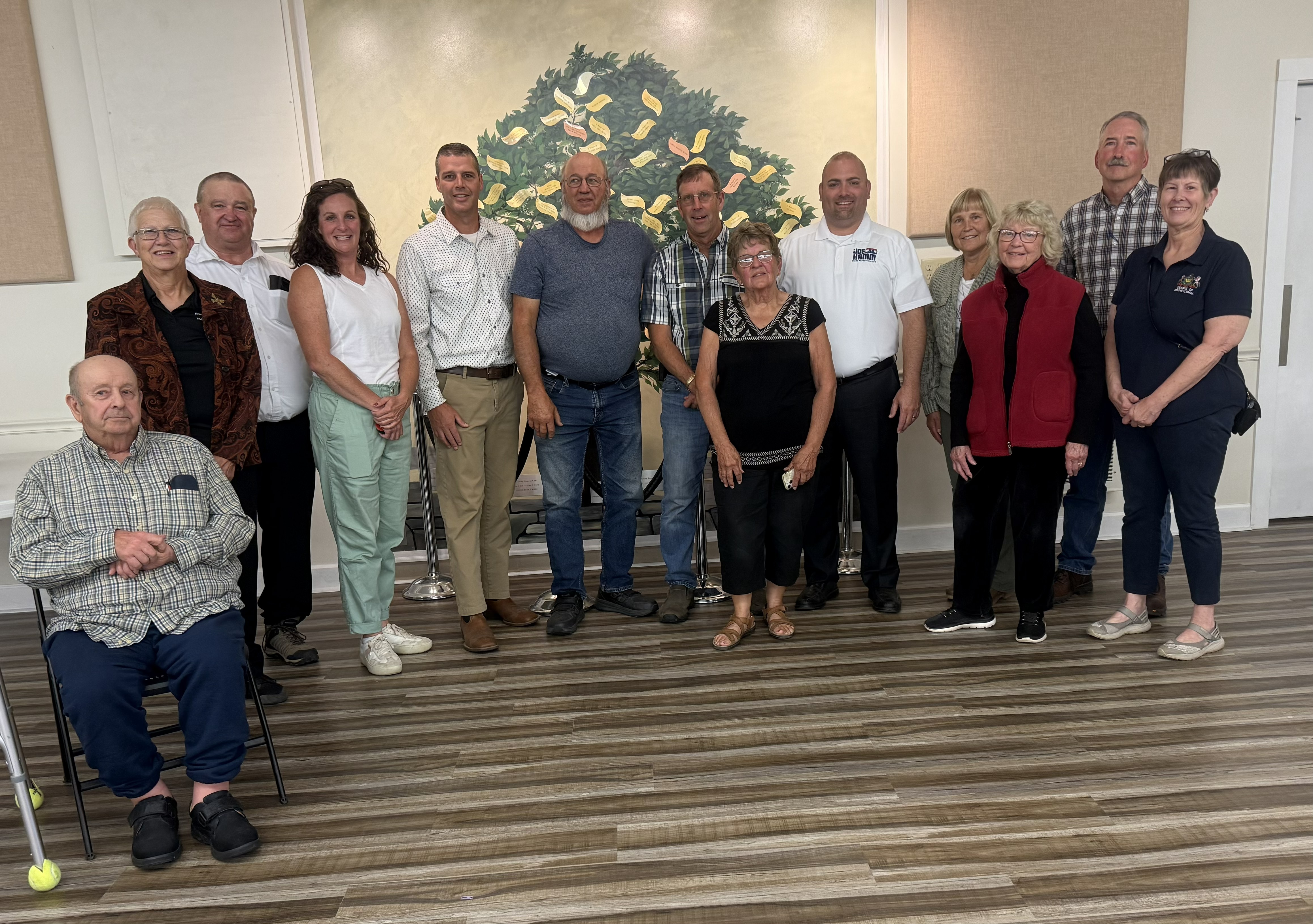A group of adults stands in a row inside a community room, smiling toward the camera. One person sits in front using a walker. A mural of a tree hangs behind the group.