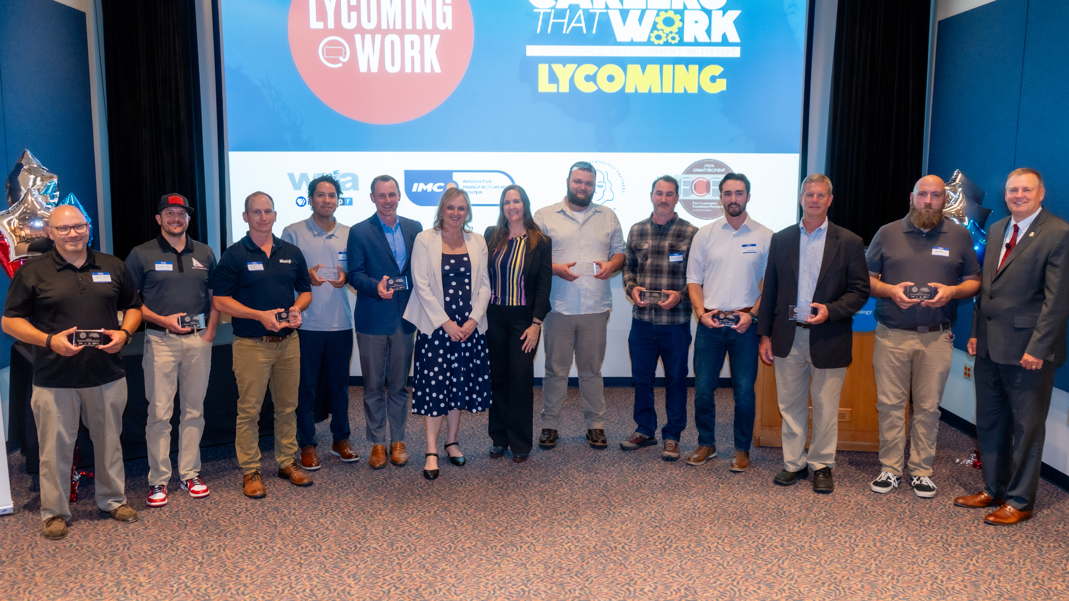 A group of people stand in a row on a stage, posing for a photo. Many hold small award plaques. Behind them is a large screen displaying logos for the Lycoming Work initiative and partner organizations. The group is smiling, and silver star‑shaped balloons decorate the sides of the stage.