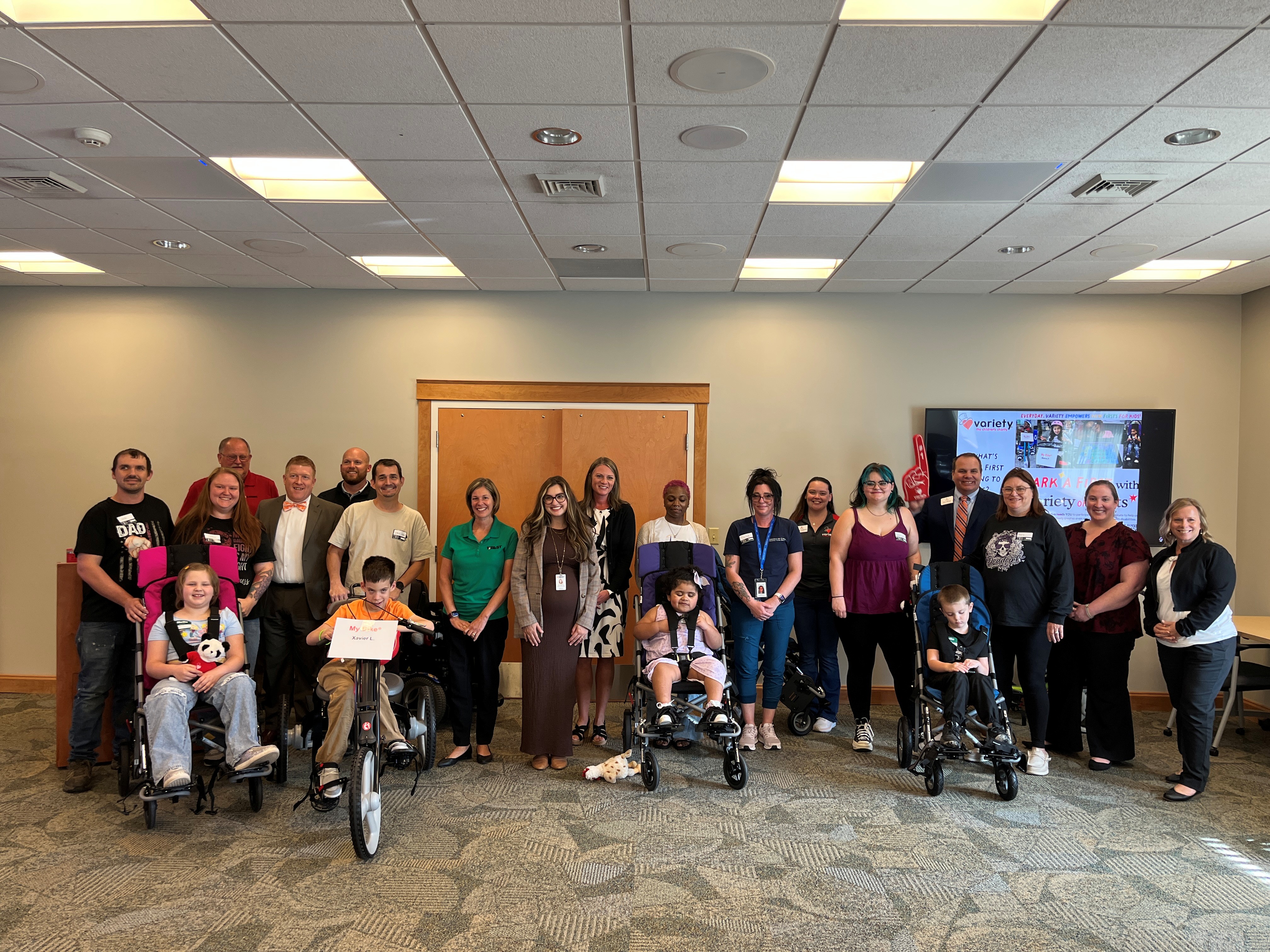 A group of children and adults pose together in a meeting room. Several children use adaptive bikes or mobility devices. A presentation screen with graphics is visible in the background.