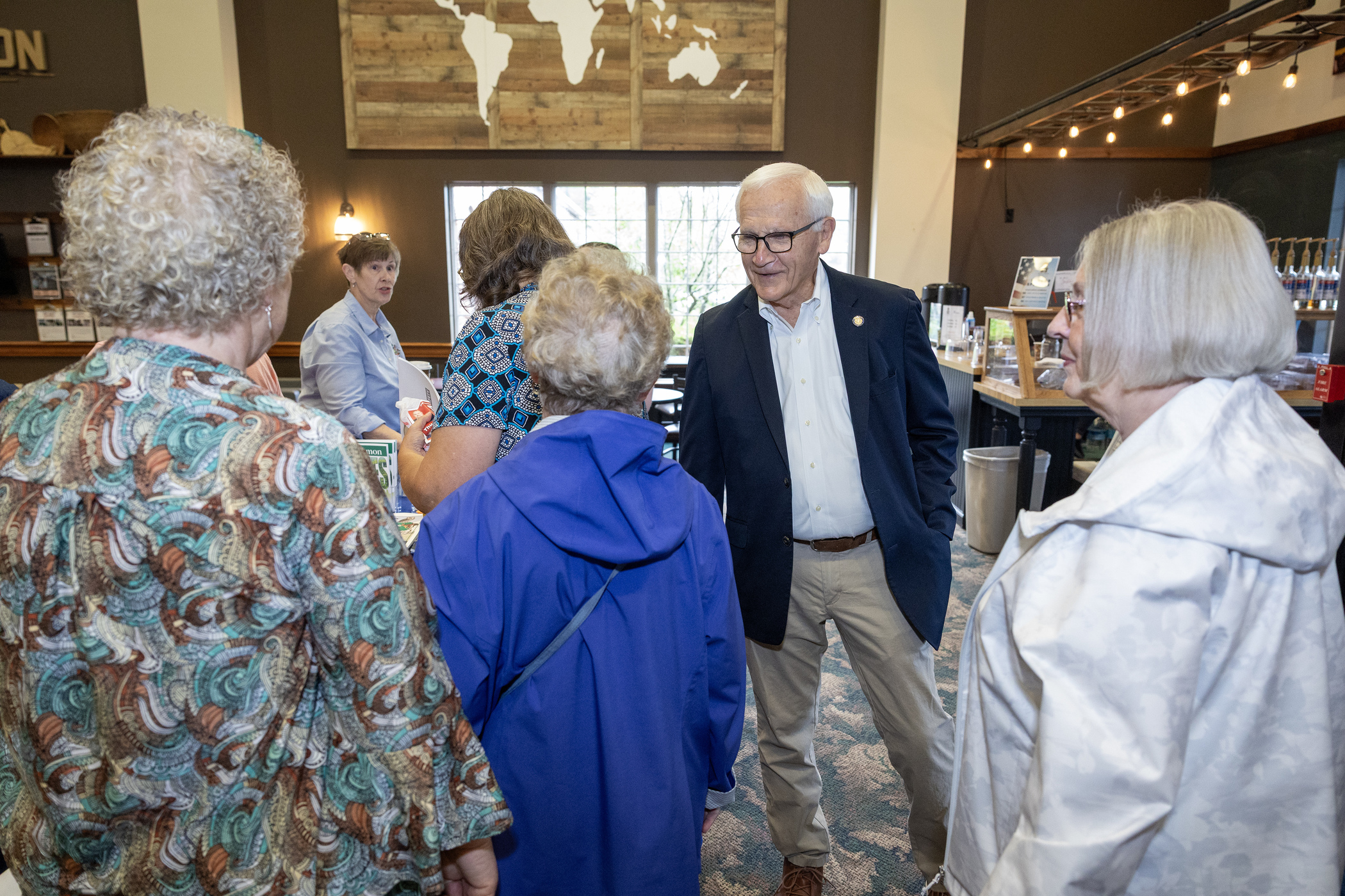 Sen. Yaw speaks with several older adults gathered around him in a brightly lit café-style space. A staff member stands behind a counter in the background.