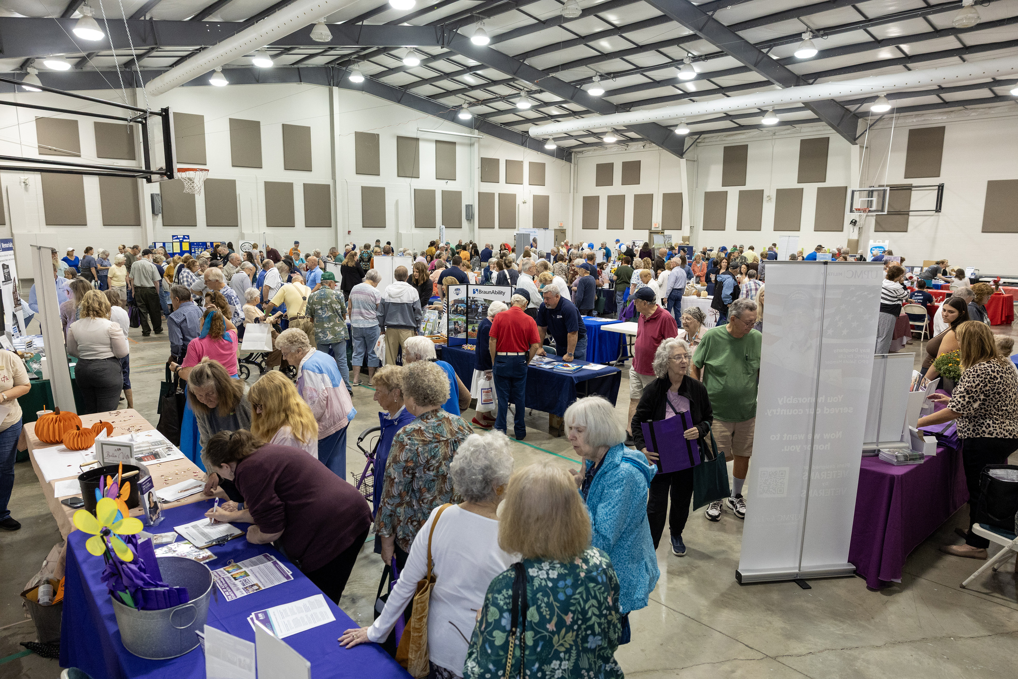 A large crowd moves through a busy indoor expo with rows of tables and informational booths. Attendees talk with vendors and browse displays throughout the open gymnasium-like space.