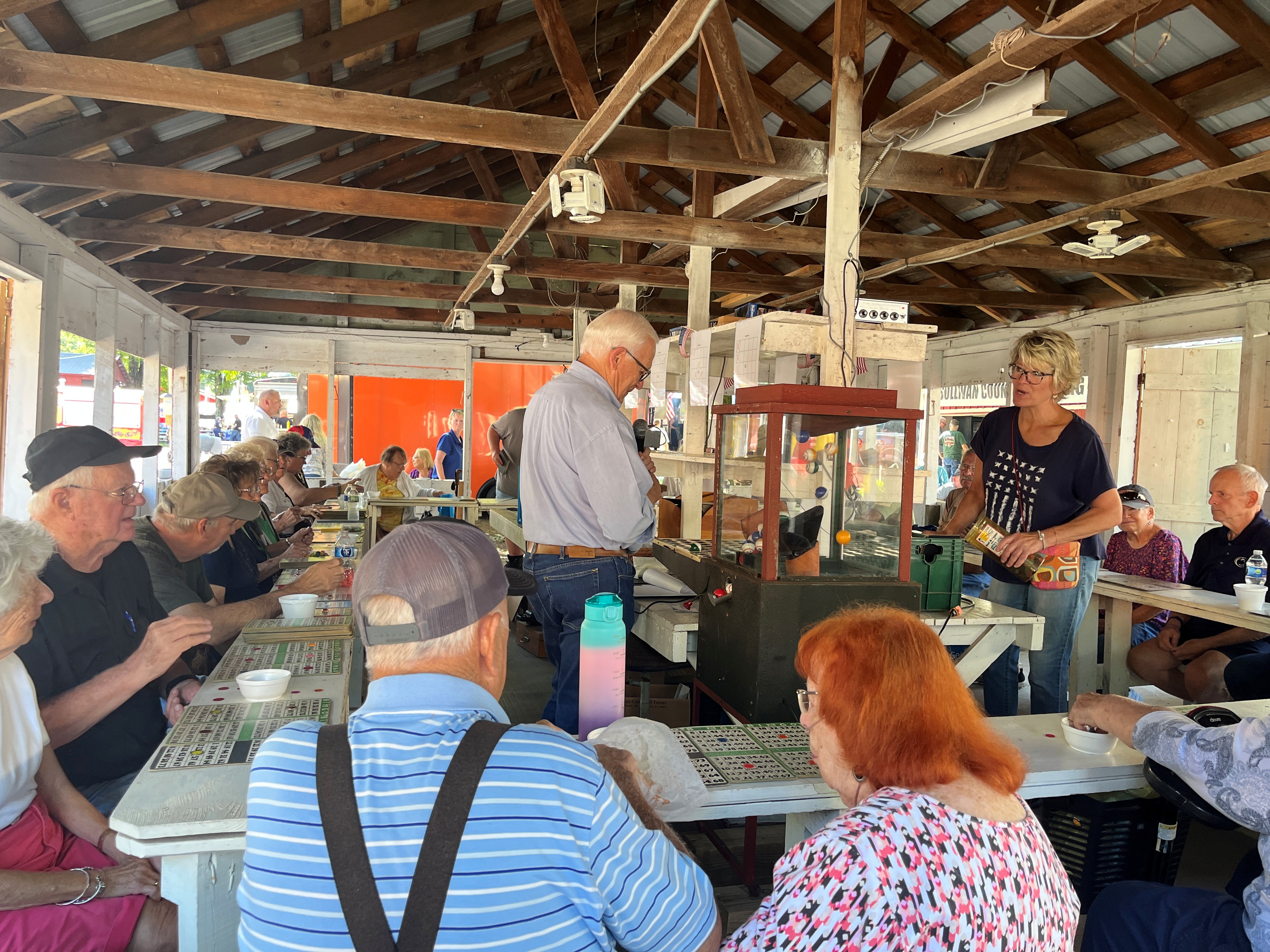 Adults seated inside an open‑air pavilion playing a table game, with cards and small bowls in front of them. Yaw stands at a central counter operating a gaming machine while others watch. The pavilion has exposed beams and simple wooden walls.
