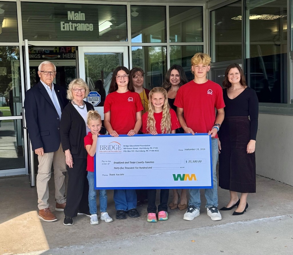 Sen. Gene Yaw and Rep. Tina Pickett, along with Waste Management officials, teachers and students stand together outside a building entrance, smiling and holding an oversized ceremonial check. Four children wearing matching red shirts stand in front, with adults gathered behind them. The check displays a donation amount of $35,500 from a foundation and a waste management company. The group is posed in front of large windows under a sign that reads “Main Entrance.”