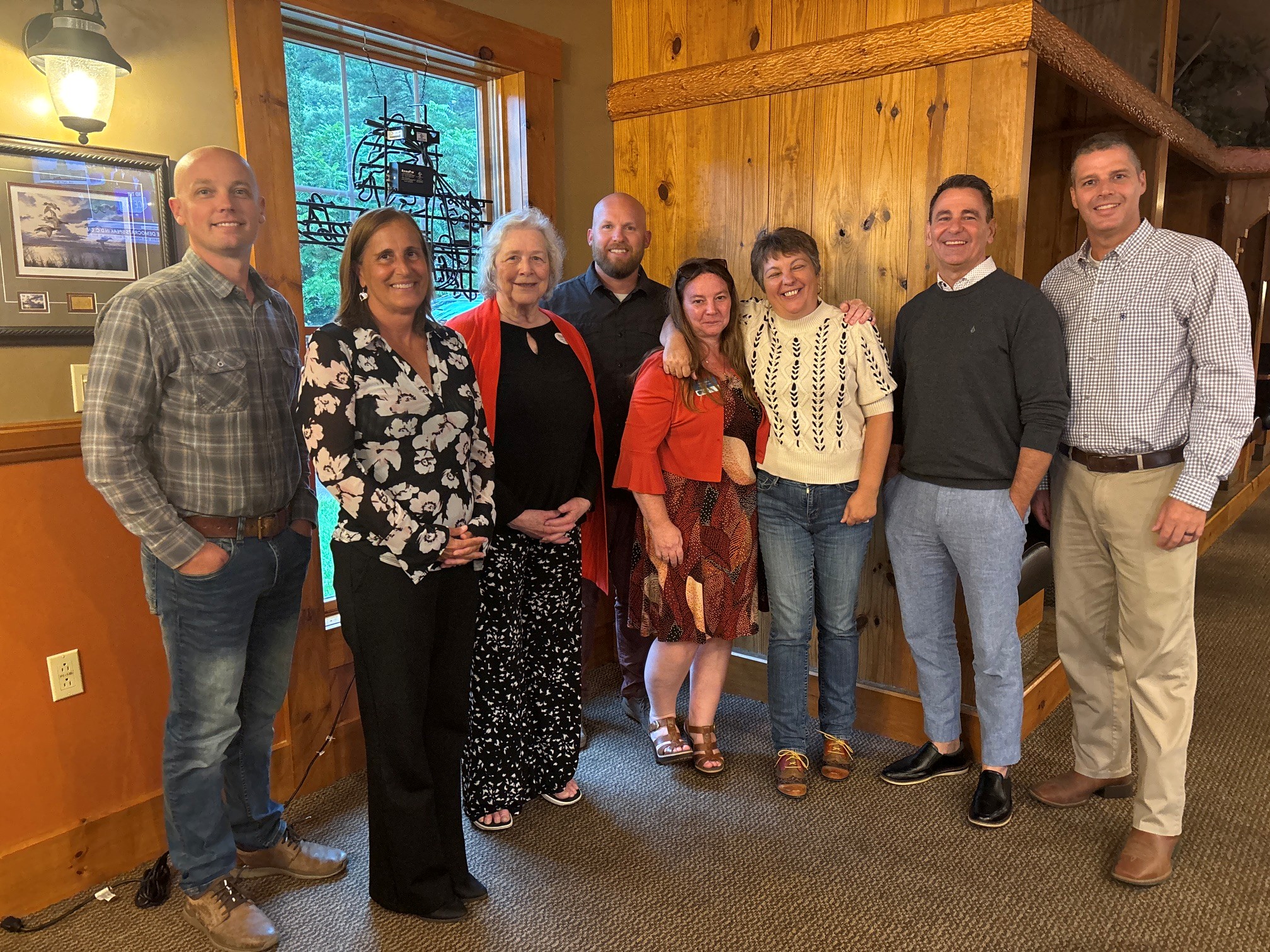 Eight adults stand side by side inside a rustic wooden room, smiling for a group photo near a window and wooden wall paneling.