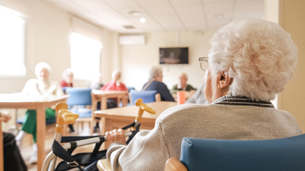 Back view of senior woman with short white hair sitting on chair with foldable walker with friends in blurred background at nursing home
