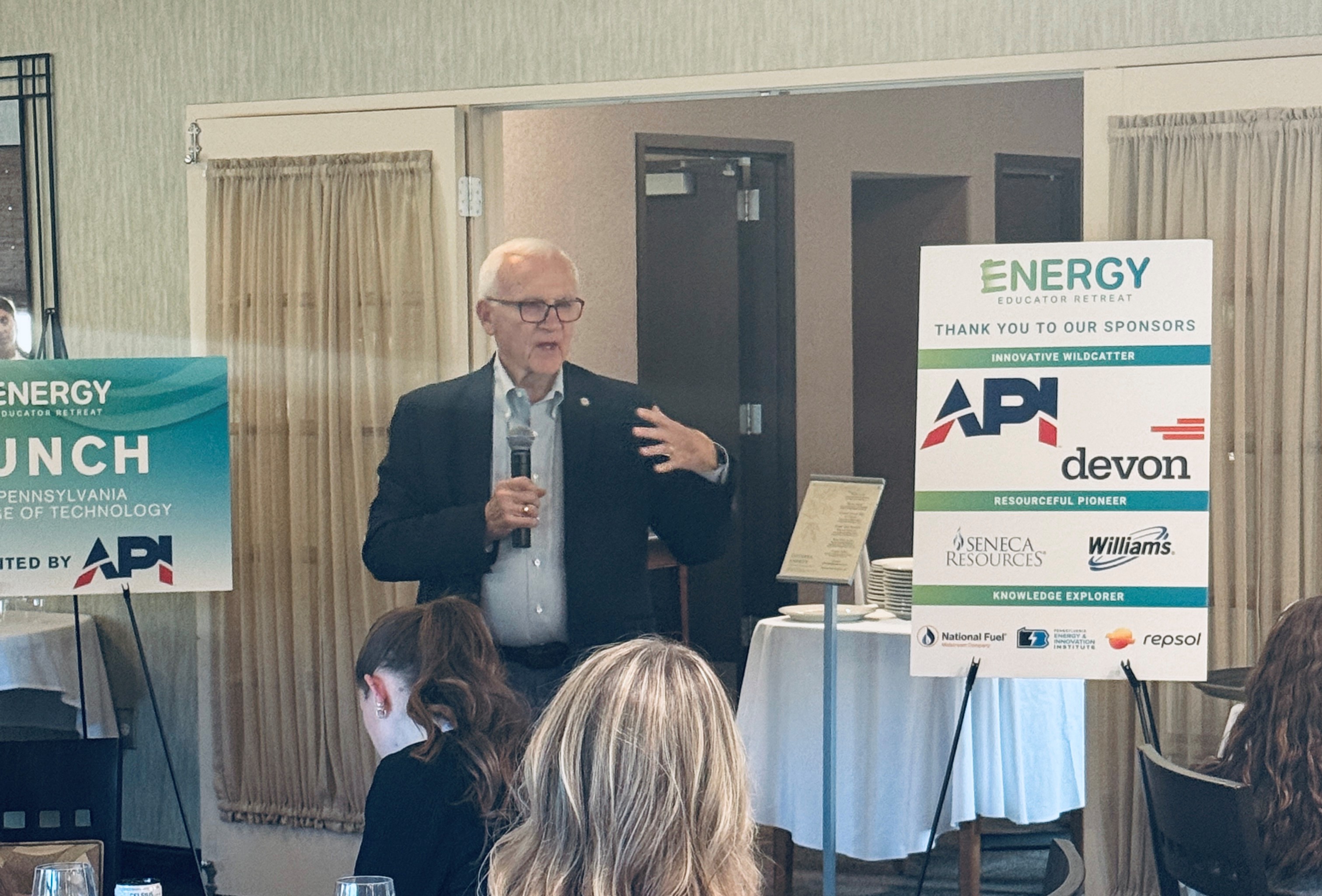 Sen. Yaw holds a microphone and addresses attendees at the Energy Educator Retreat. Sponsor signs for API, Devon, Seneca Resources, Williams, National Fuel, Pennsylvania Energy & Innovation Institute and Repsol are displayed.